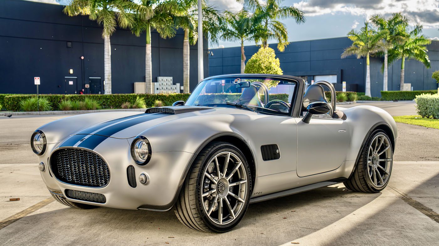 A silver sports car with black racing stripes parked in front of palm trees and a dark building.