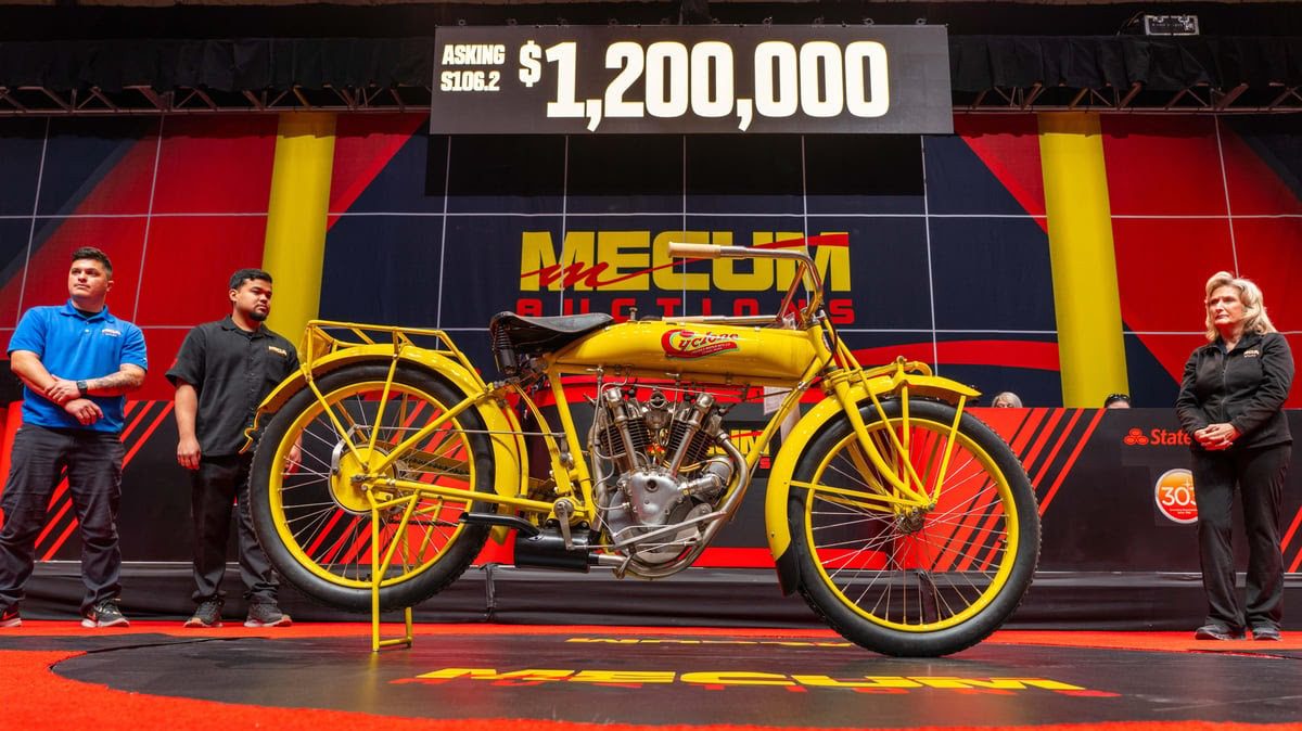 A vintage yellow motorcycle is on display at a Mecum Auction in Las Vegas. The asking price is $1,200,000. Four people stand nearby, admiring the standout piece of this exclusive motorcycle auction.
