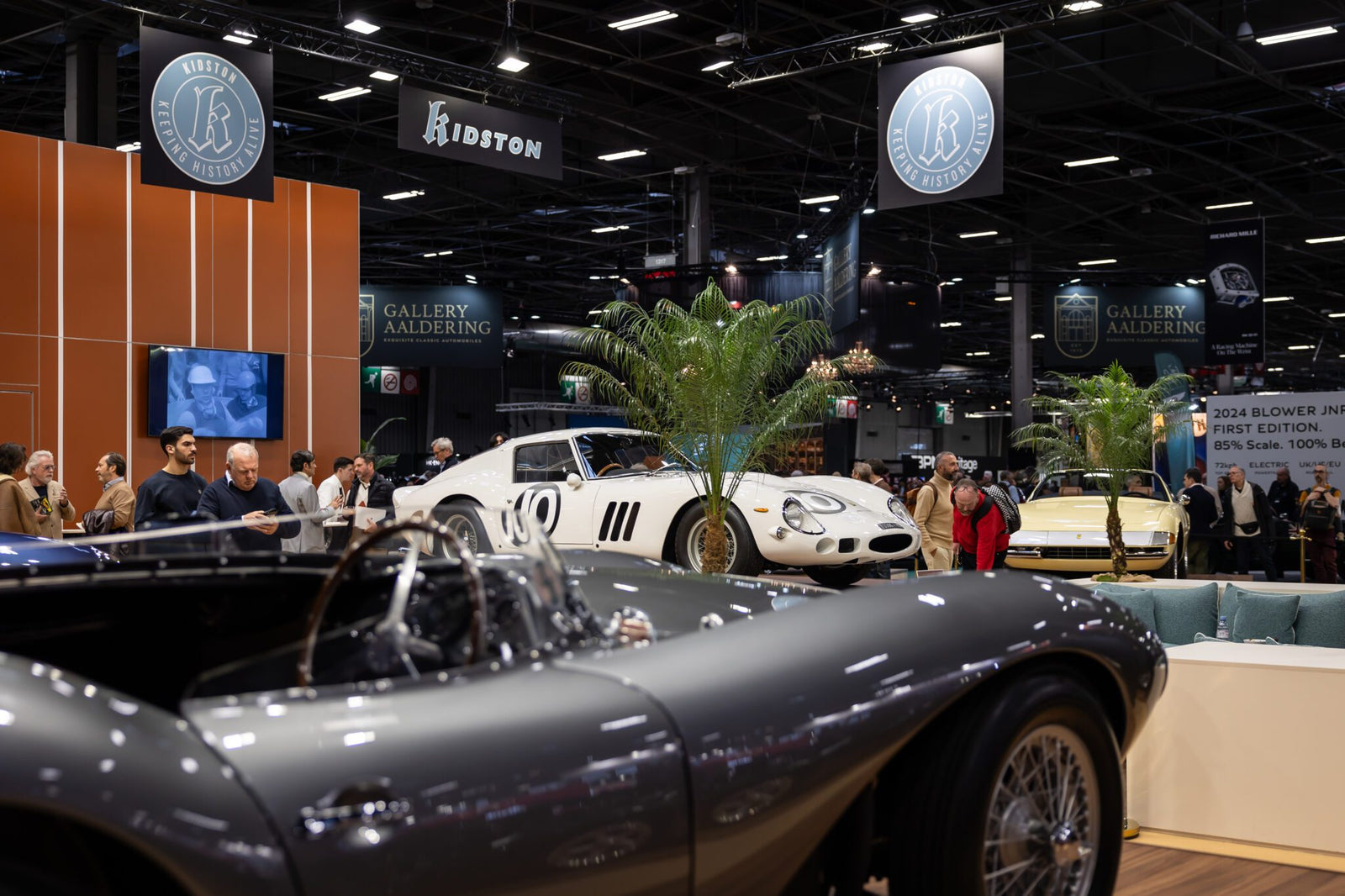 Experience European elegance at Rétromobile NY, an indoor Manhattan car show where visitors admire classic cars, including a silver convertible in the foreground and a white sports car on display in the background.