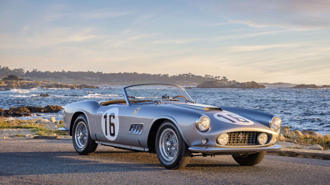 A vintage Ferrari sports car, reminiscent of the classic California Spider, proudly displays the number 16 as it rests by the ocean under a partly cloudy sky, with Amelia Island's rocky coastline painting a picturesque background.