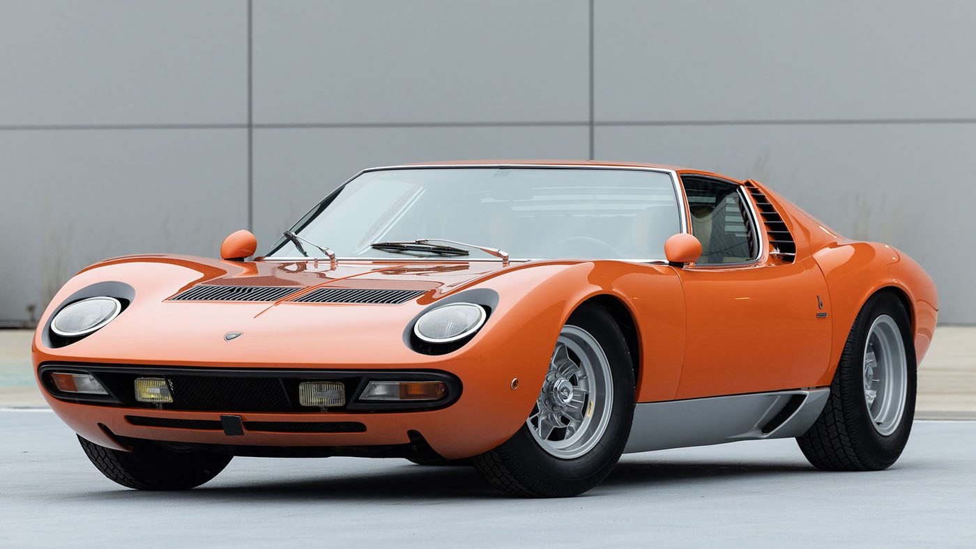 A 1971 Lamborghini Miura, with its orange vintage allure and sleek design, is poised at an angle on the gray surface. This classic beauty could easily attract attention at prestigious events like Broad Arrow Auctions or Amelia Island.