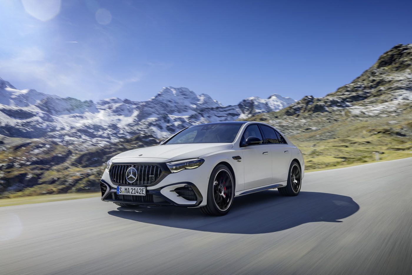A sleek Mercedes-AMG E 53 hybrid car glides along a mountain road, framed by snowy peaks under a clear blue sky, offering a glimpse into the future of 2025 motoring.