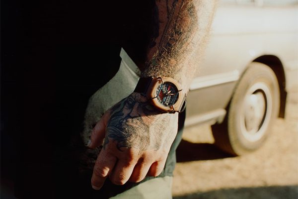 A tattooed person wearing a brown Desert King Watch stands beside a beige car, with sunlight casting shadows on their hand and arm.