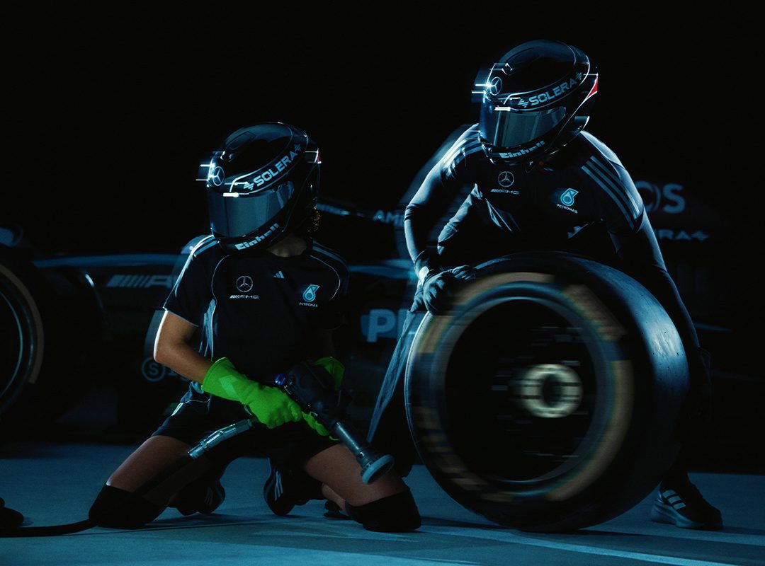 Two pit crew members in racing gear, one holding a tire and the other using a tool, perform a pit stop under dim lighting.
