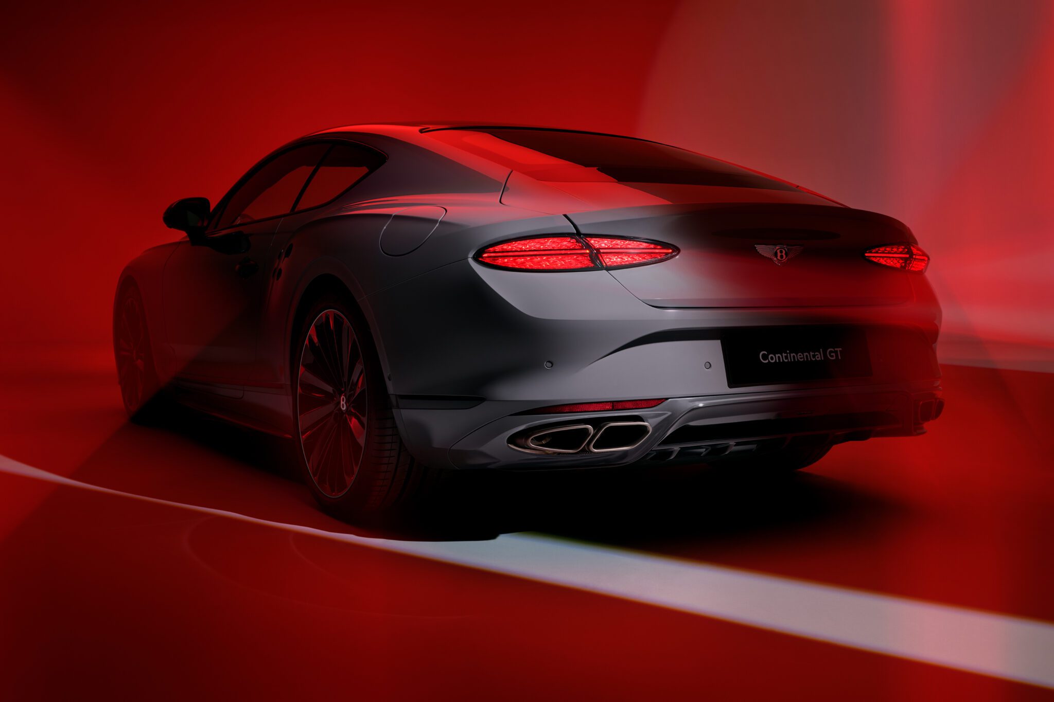 A sleek, silver Bentley Continental GT sports coupe viewed from the rear, illuminated by dramatic red lighting and shadows, showcases its unmistakable exhaust note—a testament to the Akrapovič partnership with Bentley Motors.