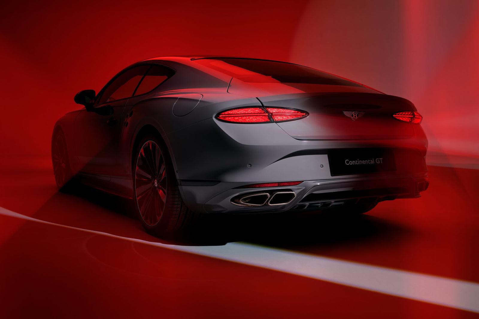 A sleek, silver Bentley Continental GT sports coupe viewed from the rear, illuminated by dramatic red lighting and shadows, showcases its unmistakable exhaust note—a testament to the Akrapovič partnership with Bentley Motors.