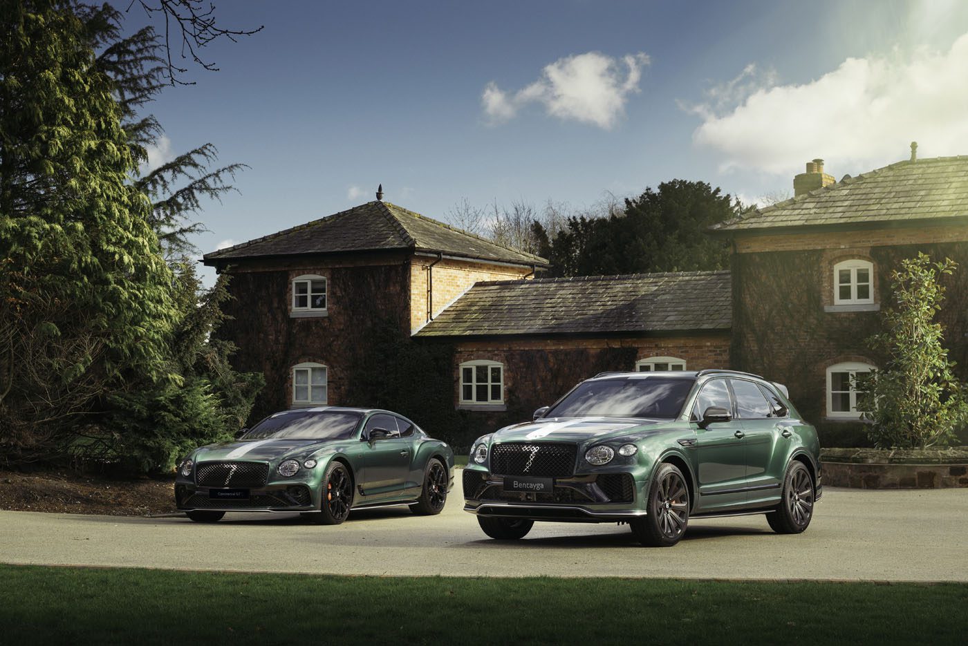 Two green luxury cars from the Le Mans Collection are parked on a driveway in front of a rustic house, surrounded by trees under a partly cloudy sky.