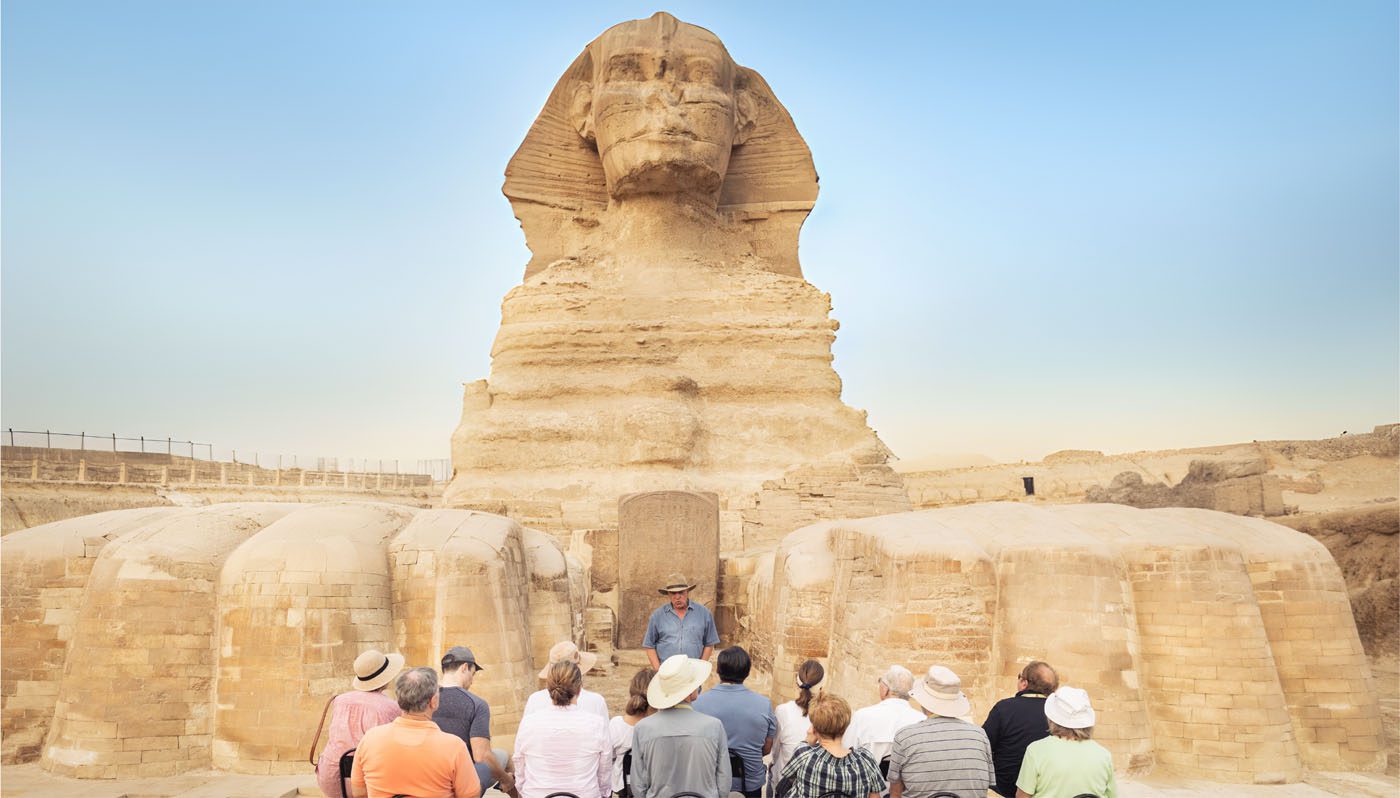 A group of people enjoys a Luxury Travel experience, attentively listening to a guide in front of the Great Sphinx of Giza under a clear blue sky.