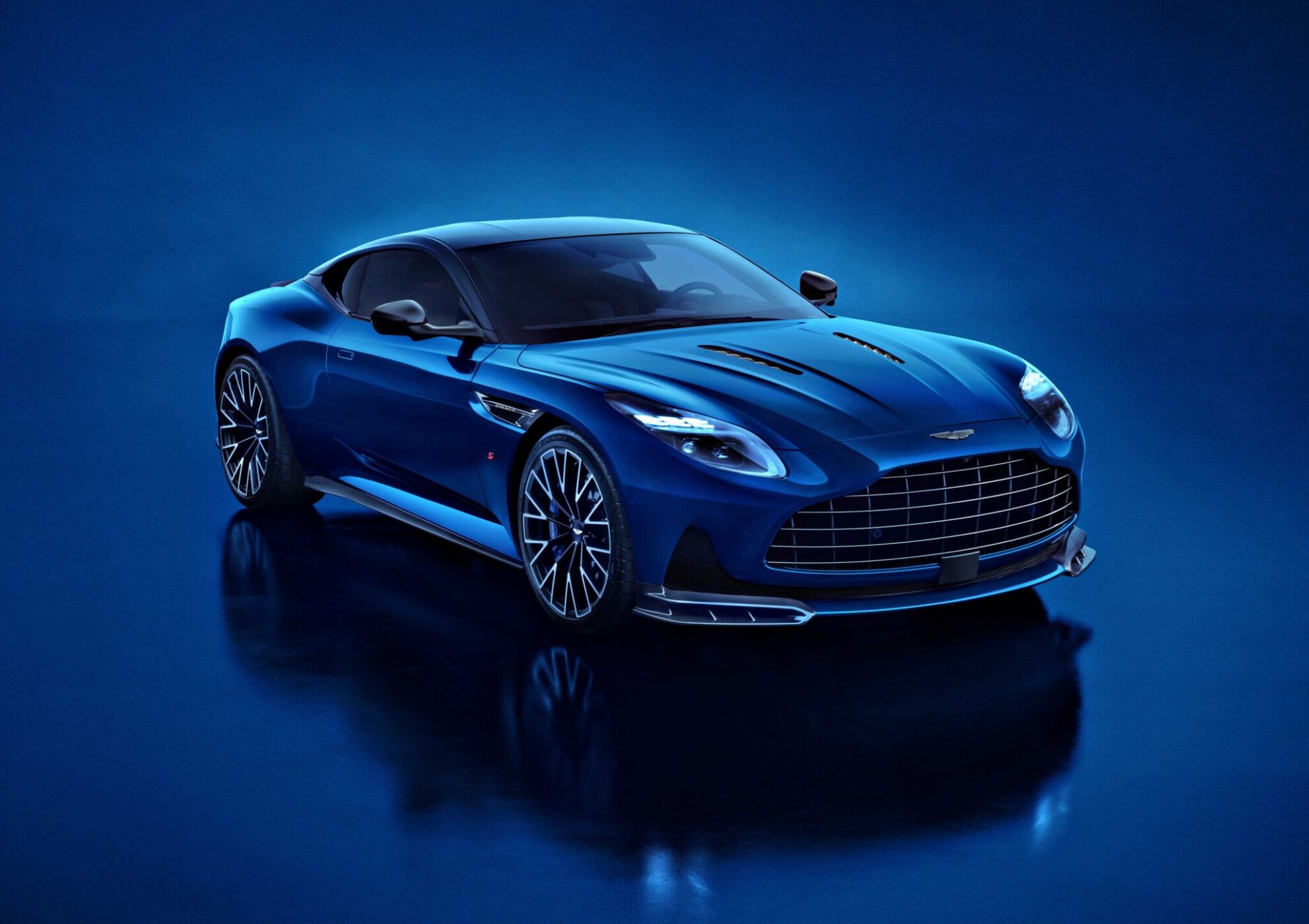 A blue Aston Martin DB12 S luxury sports car is parked on a glossy blue surface, photographed from the front left angle under bright lighting—an exquisite addition to the Super Tourers class.