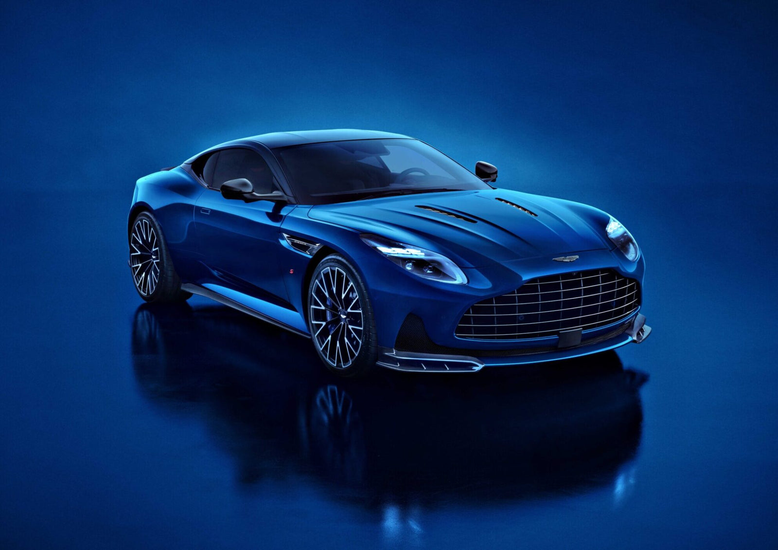 A blue Aston Martin DB12 S luxury sports car is parked on a glossy blue surface, photographed from the front left angle under bright lighting—an exquisite addition to the Super Tourers class.