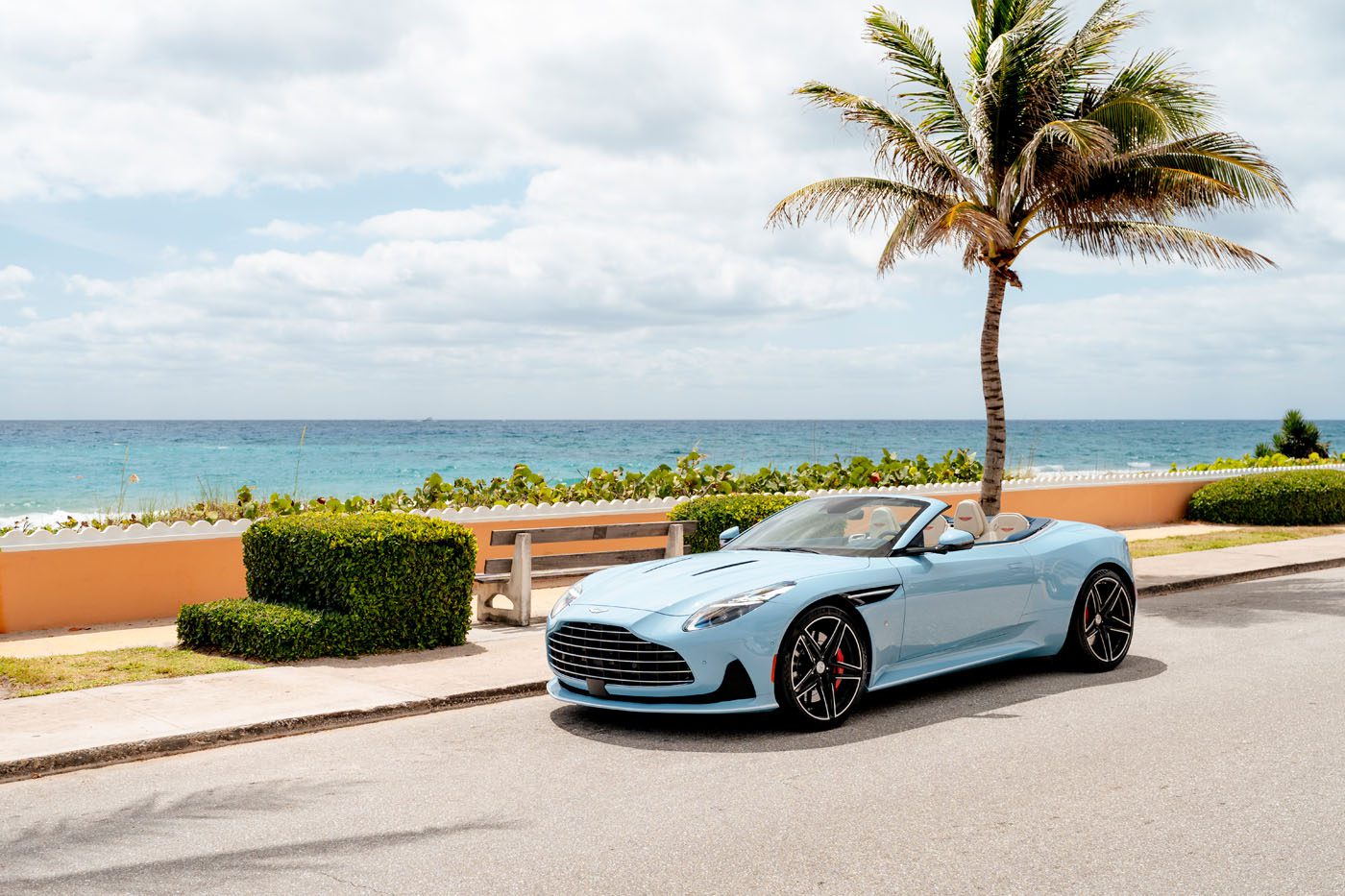 A light blue Aston Martin DB12 Volante convertible is parked on a coastal road near a palm tree, with the ocean and a partly cloudy sky in the background—capturing the essence of the exclusive Palm Beach Edition.