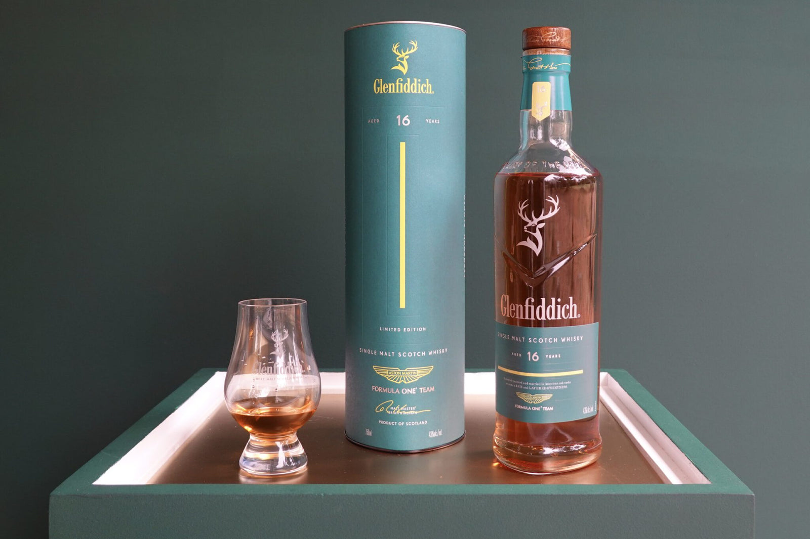 A bottle of Glenfiddich 16-year-old single malt Scotch whisky, inspired by Aston Martin’s new Formula 1 Team Scotch Whisky, with its cylindrical box and a glass of whisky on a white tray against a green background.