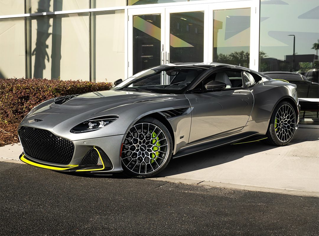 A silver sports car with neon yellow accents is parked in front of a modern glass building on a sunny day, featured in our Dealer Spotlight from Aston Martin Orlando, where exotics for sale redefine luxury and performance.