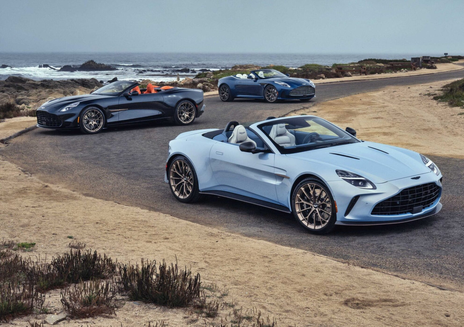 Three convertible sports cars from the Thrillseeker Collection drive along a coastal road, with the ocean and rocky shoreline in the background.