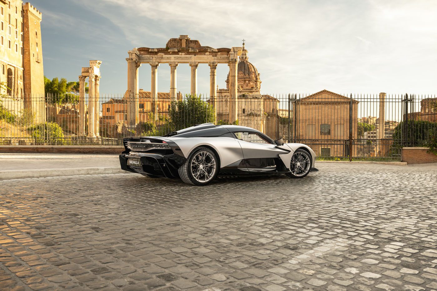 A silver sports car is parked on a cobblestone street in front of ancient Roman ruins under a partly cloudy sky.