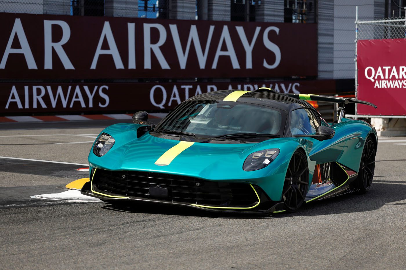 A teal Aston Martin sports car with yellow accents drives on a racetrack, with Qatar Airways banners visible in the background, capturing the thrill of a Monaco Grand Prix moment.