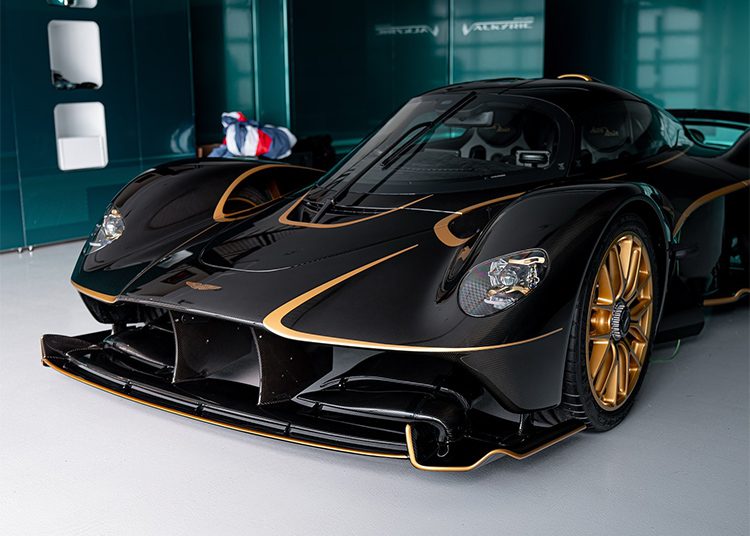 A black Aston Martin Valkyrie with aerodynamic design, exposed carbon accents, and 24K gold wheels is parked indoors on a light-colored floor.