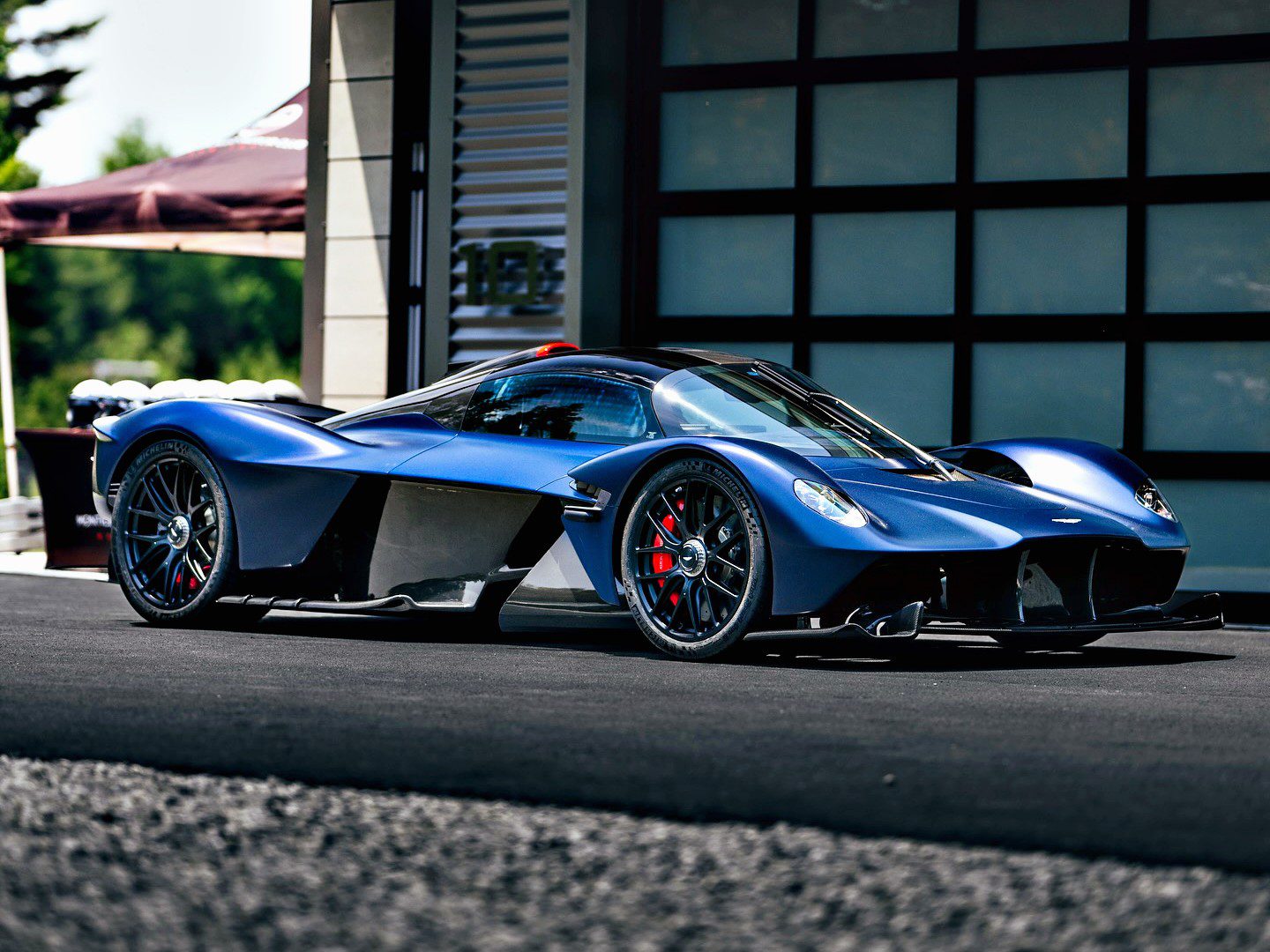 A Satin Mariana Blue Aston Martin Valkyrie hypercar is parked on a paved surface in front of a modern building.
