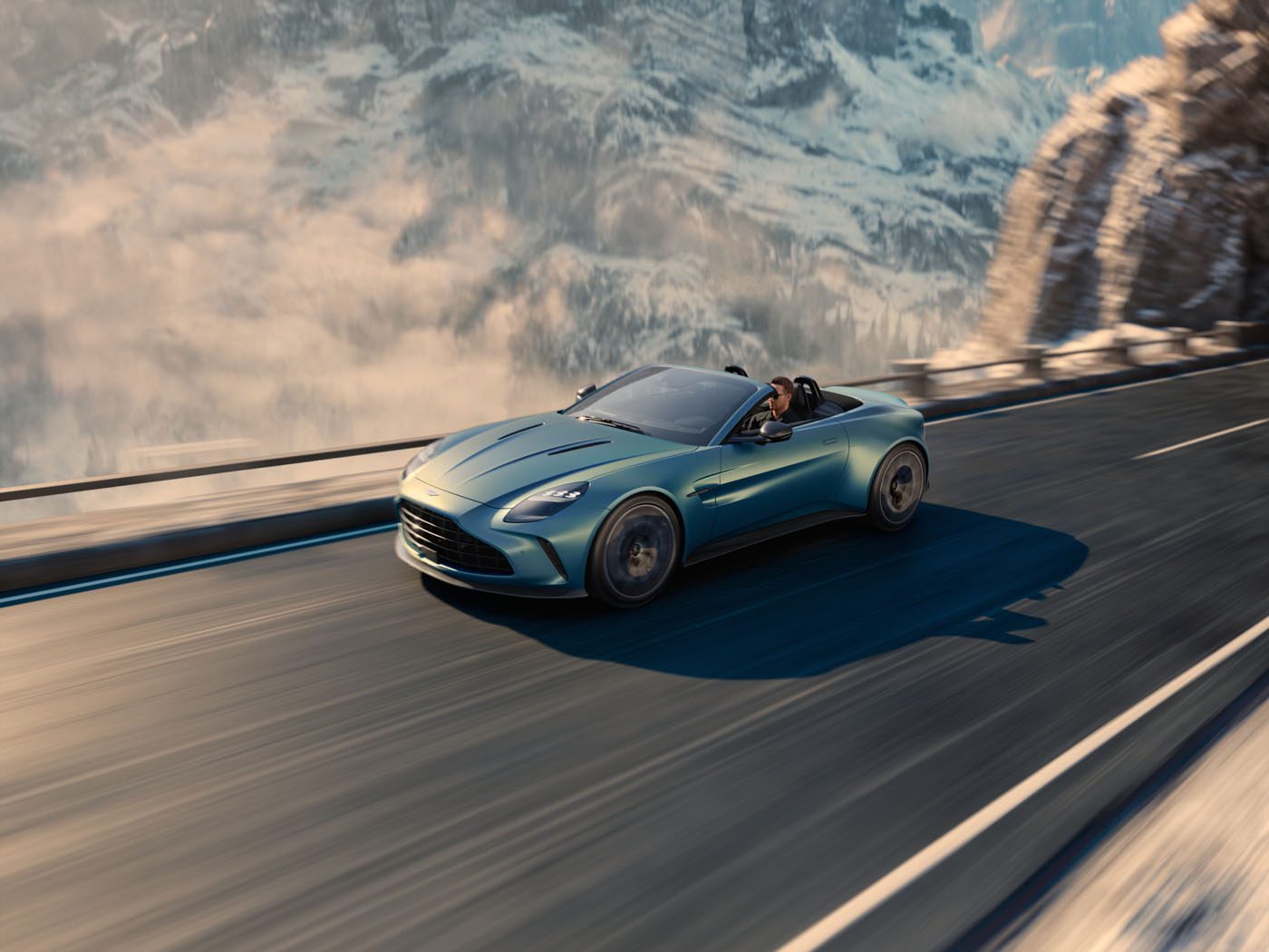 A sleek, blue Aston Martin Vantage Roadster gracefully drives on a mountainous road with snow-covered peaks in the background, capturing the thrill of adventure and the elegance of a new car release.