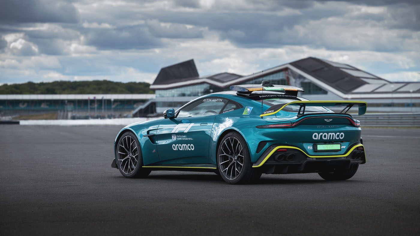 A teal Aston Martin Vantage S Formula 1 Safety Car with Aramco branding is parked on a racetrack, set against modern buildings and a cloudy sky in the background.