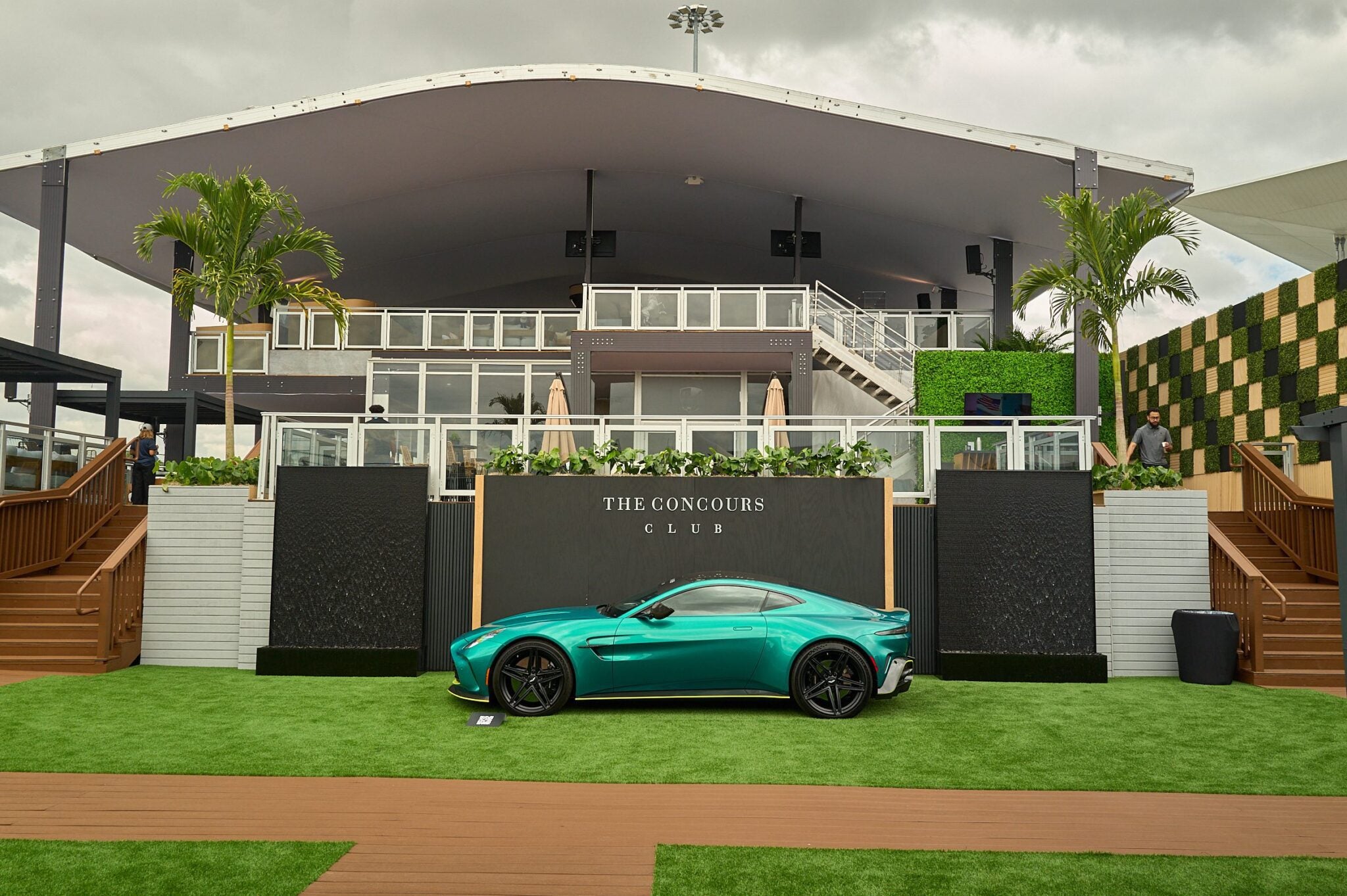 A turquoise sports car is parked on green artificial grass in front of a modern building labeled "The Concours Club," with palm trees and cloudy skies in the background.