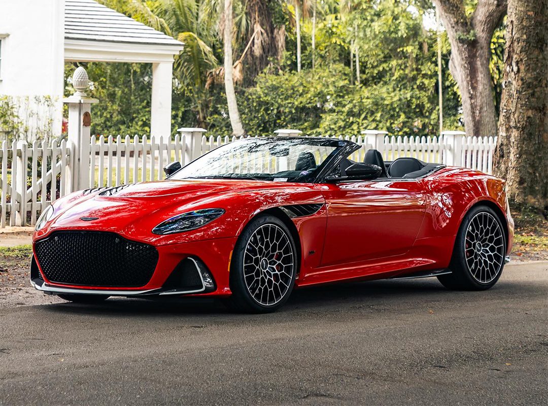 A red sports car from The Collection is parked on a shaded street, flanked by a white picket fence and trees in the background, reminiscent of classic Aston Martins.