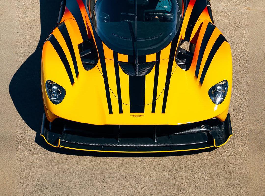 An Aston Martin Valkyrie, valued at $4M, is seen from above. This yellow sports car with sleek black racing stripes rests trackside on a concrete surface.
