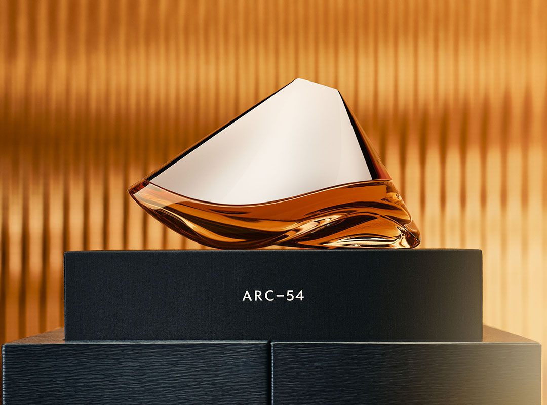 A sleek, geometrically shaped glass bottle of Arc-54 perfume sits on a black box against a gold, textured background, reminiscent of the elegance and precision of an Aston Martin Valkyrie Hypercar.