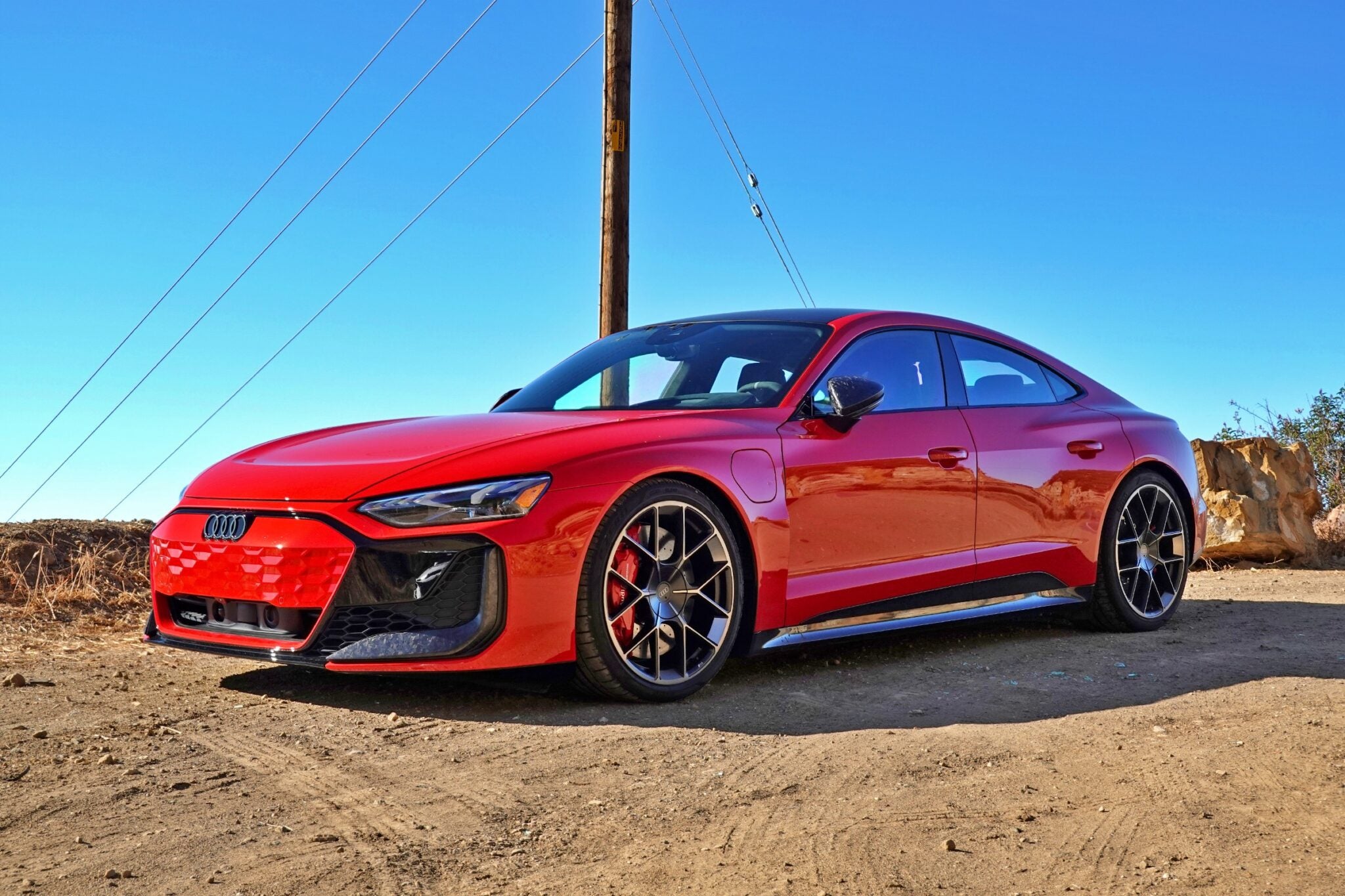 A red 2025 Audi RS e-tron GT Performance electric sports car is parked on a dirt road under a clear blue sky, with utility poles and wires in the background.
