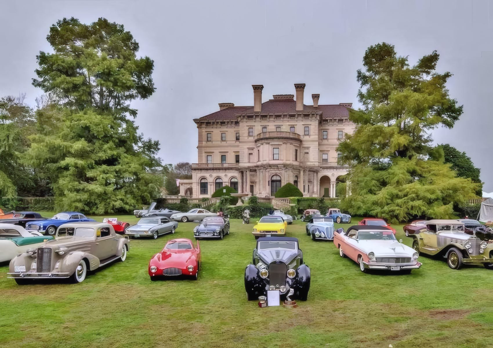 A variety of classic cars are displayed on a lawn in front of a large historic mansion on a cloudy day, capturing the spirit of East Coast Elegance: Audrain Newport Concours & Motor Week 2025.