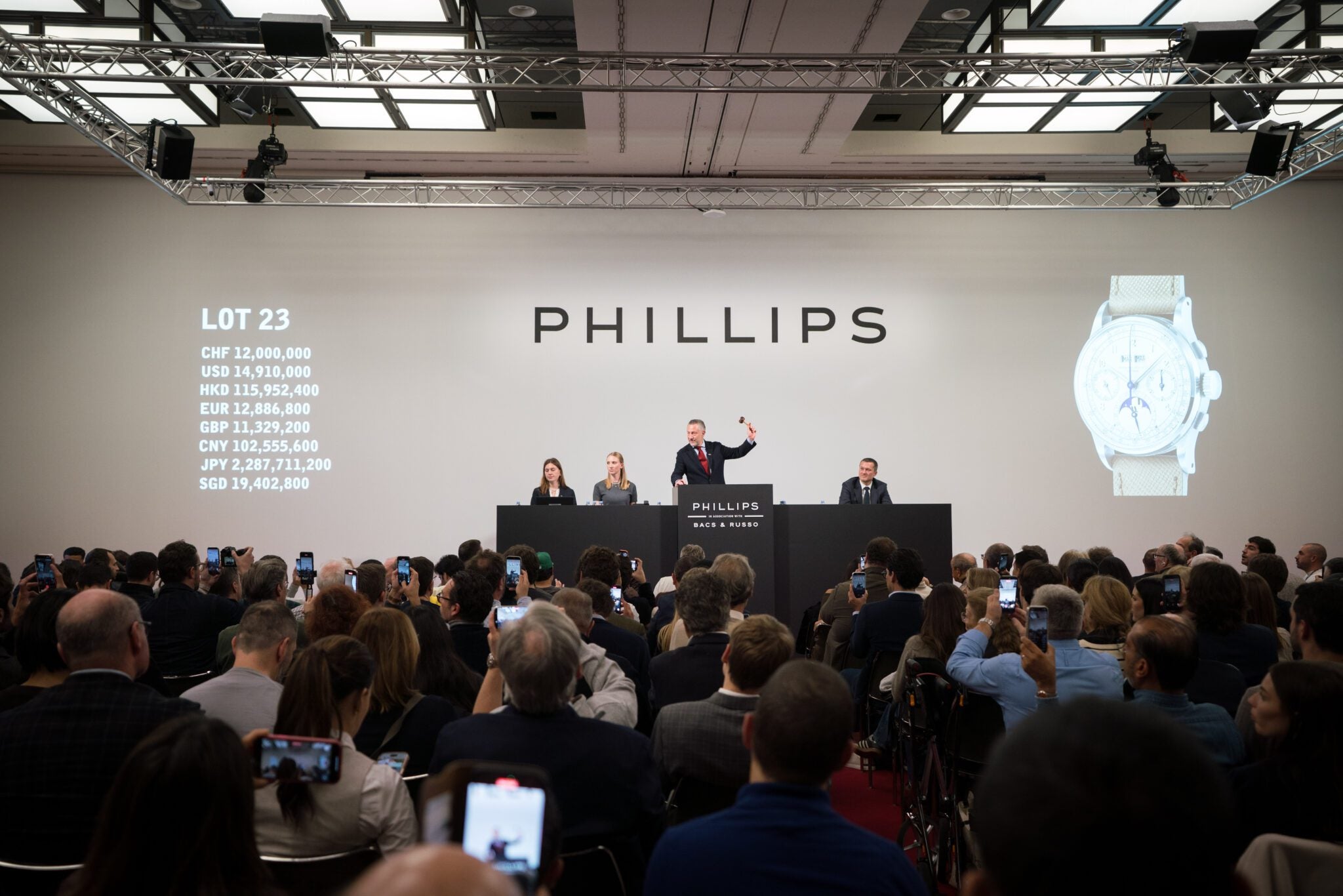 An auctioneer conducts a watch auction at Phillips, with attendees photographing the event. Currency values and an image of one of the most expensive watches are displayed on large screens during this record-breaking year.