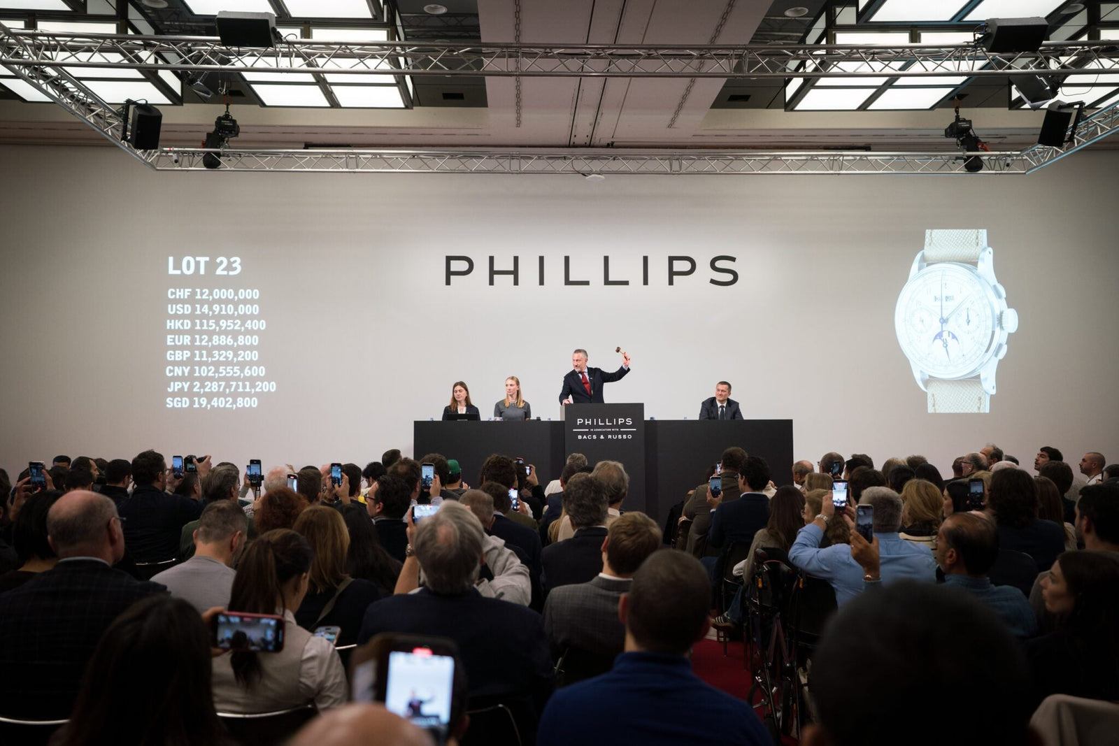 An auctioneer conducts a watch auction at Phillips, with attendees photographing the event. Currency values and an image of one of the most expensive watches are displayed on large screens during this record-breaking year.