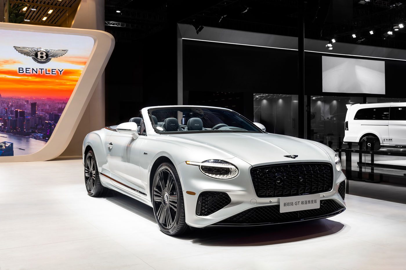 A white Bentley Continental convertible is displayed in a showroom with a Bentley sign and other vehicles in the background.