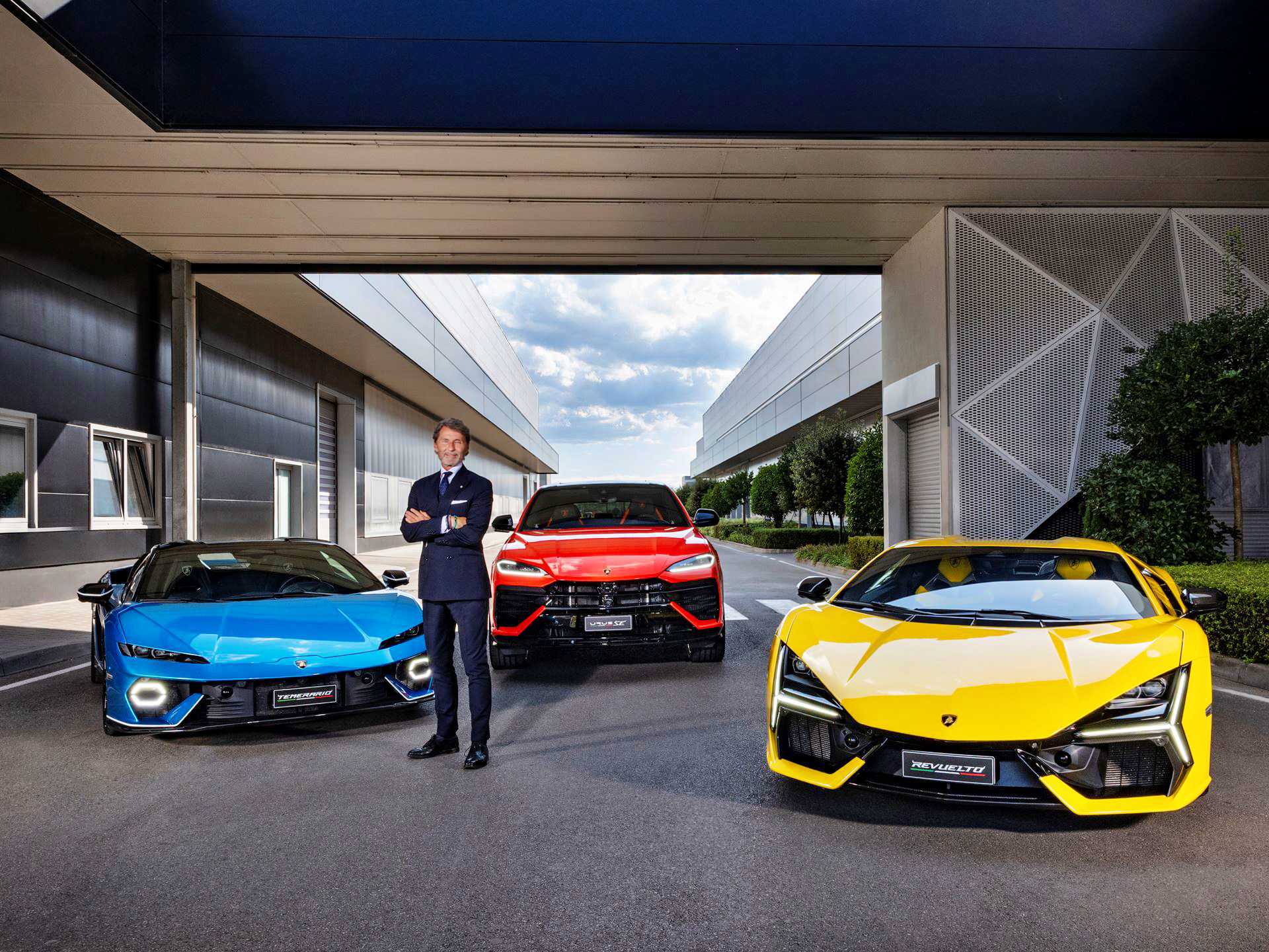 A man in a suit stands with arms crossed between a blue car, a red SUV, and a yellow sports car—reminiscent of Lamborghini’s strong Q3 results and record margins in the hybrid era—parked outside a modern building.