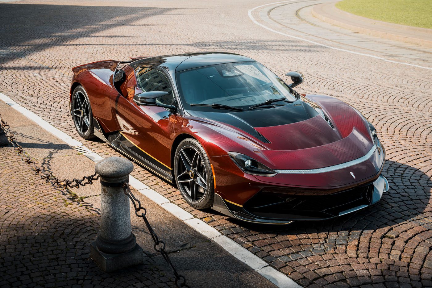 A sleek red and black Pininfarina Battista sports car is parked on a cobblestone street near a curb lined with short metal posts and chains.