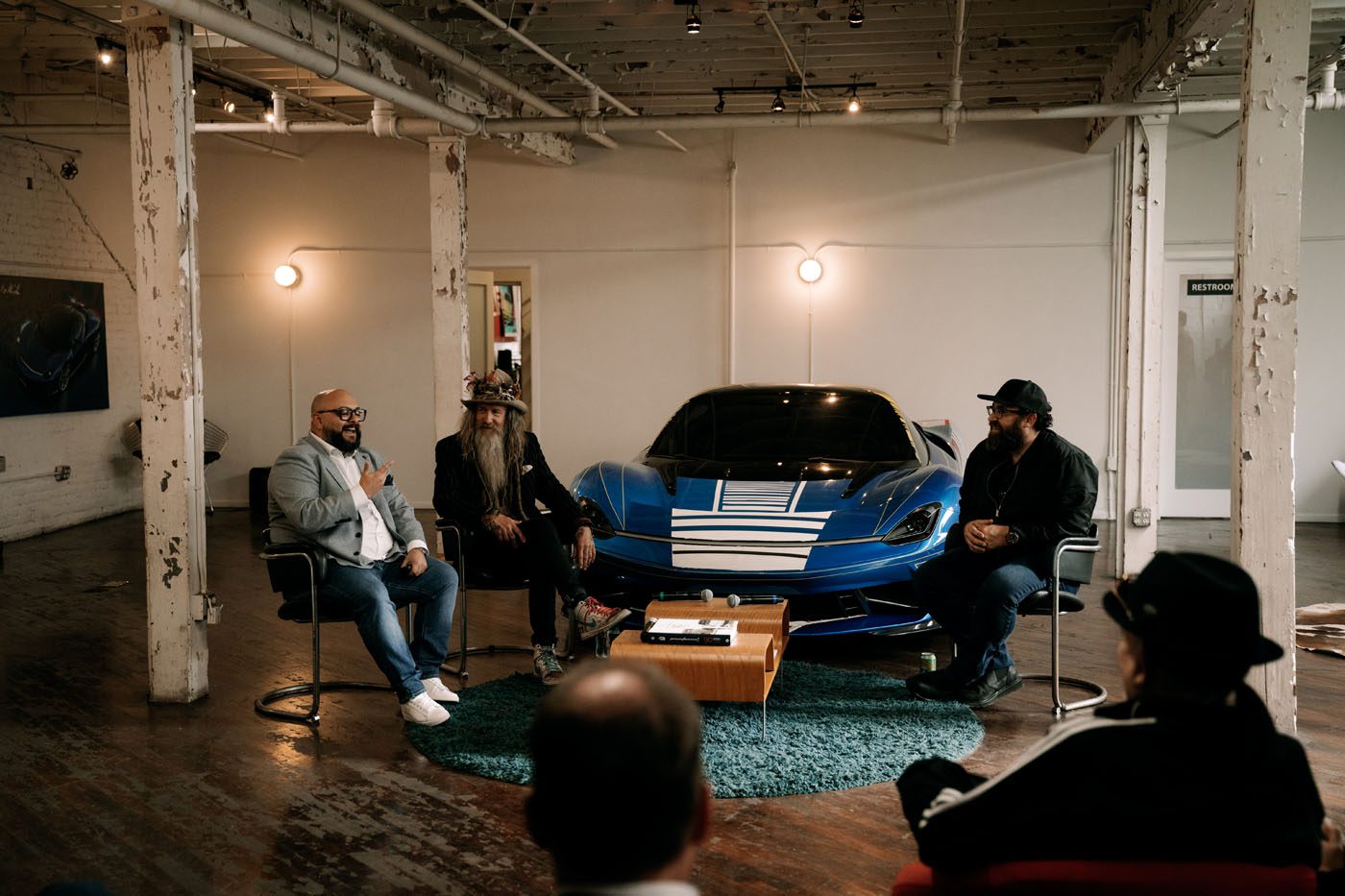 Three men sit and talk in front of a blue sports car in an industrial-style room with exposed beams and wooden floors, while a few people watch.