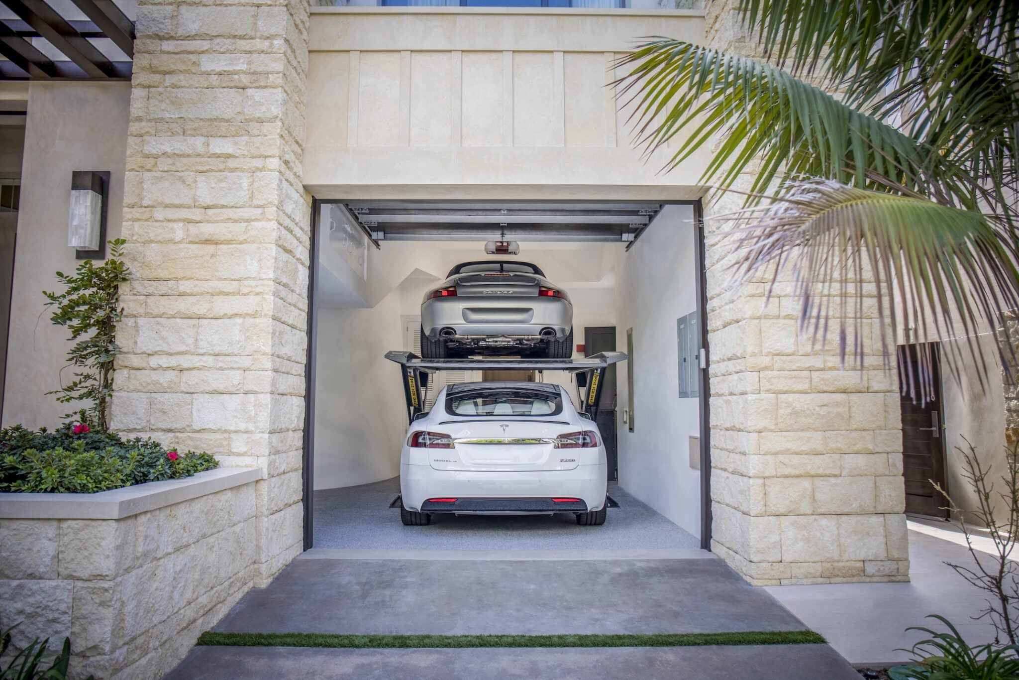 Two cars are parked in a double garage with an Autostacker car lift; a white Tesla sits below while a silver car is elevated above, maximizing garage space.