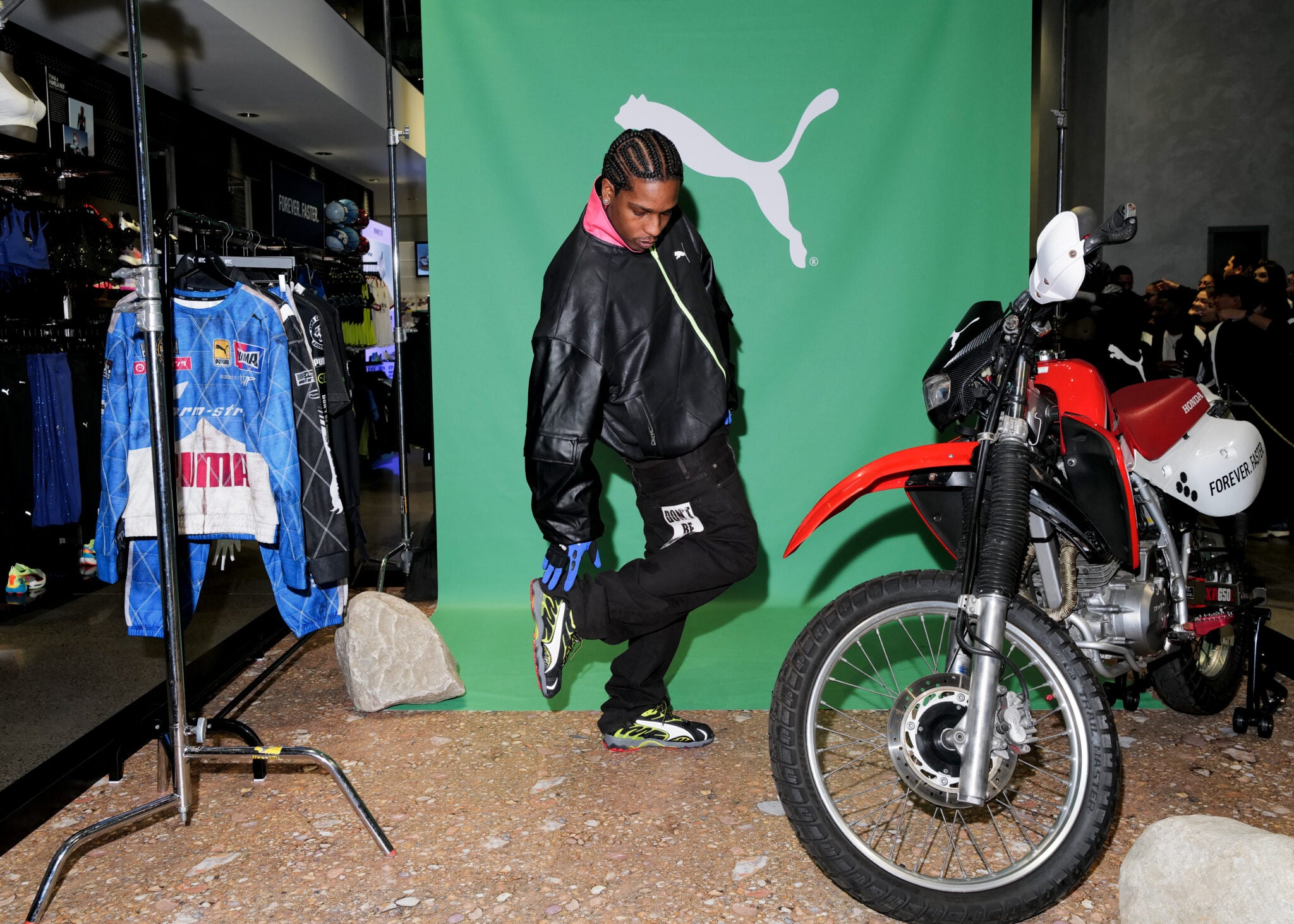A person in a black jacket poses in front of a green Puma Motorsport backdrop, alongside A$AP Rocky's new collection display, next to a red and white motorcycle and a clothing rack.