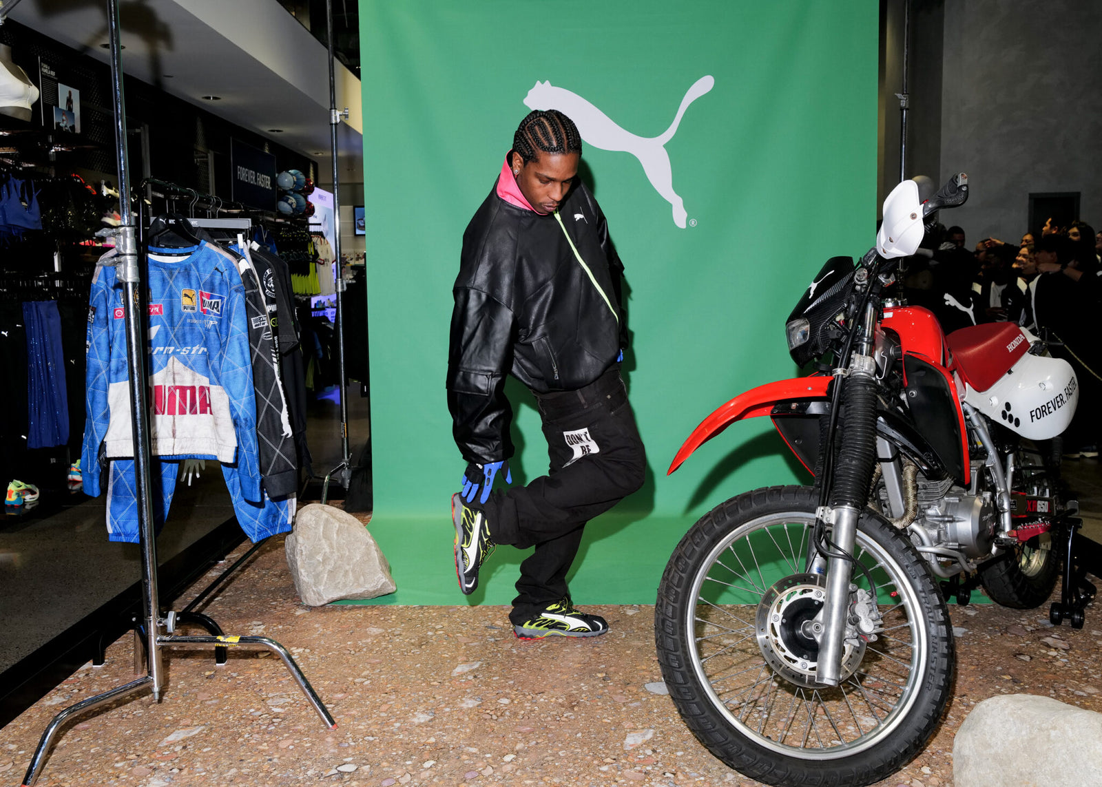 A person in a black jacket poses in front of a green Puma Motorsport backdrop, alongside A$AP Rocky's new collection display, next to a red and white motorcycle and a clothing rack.
