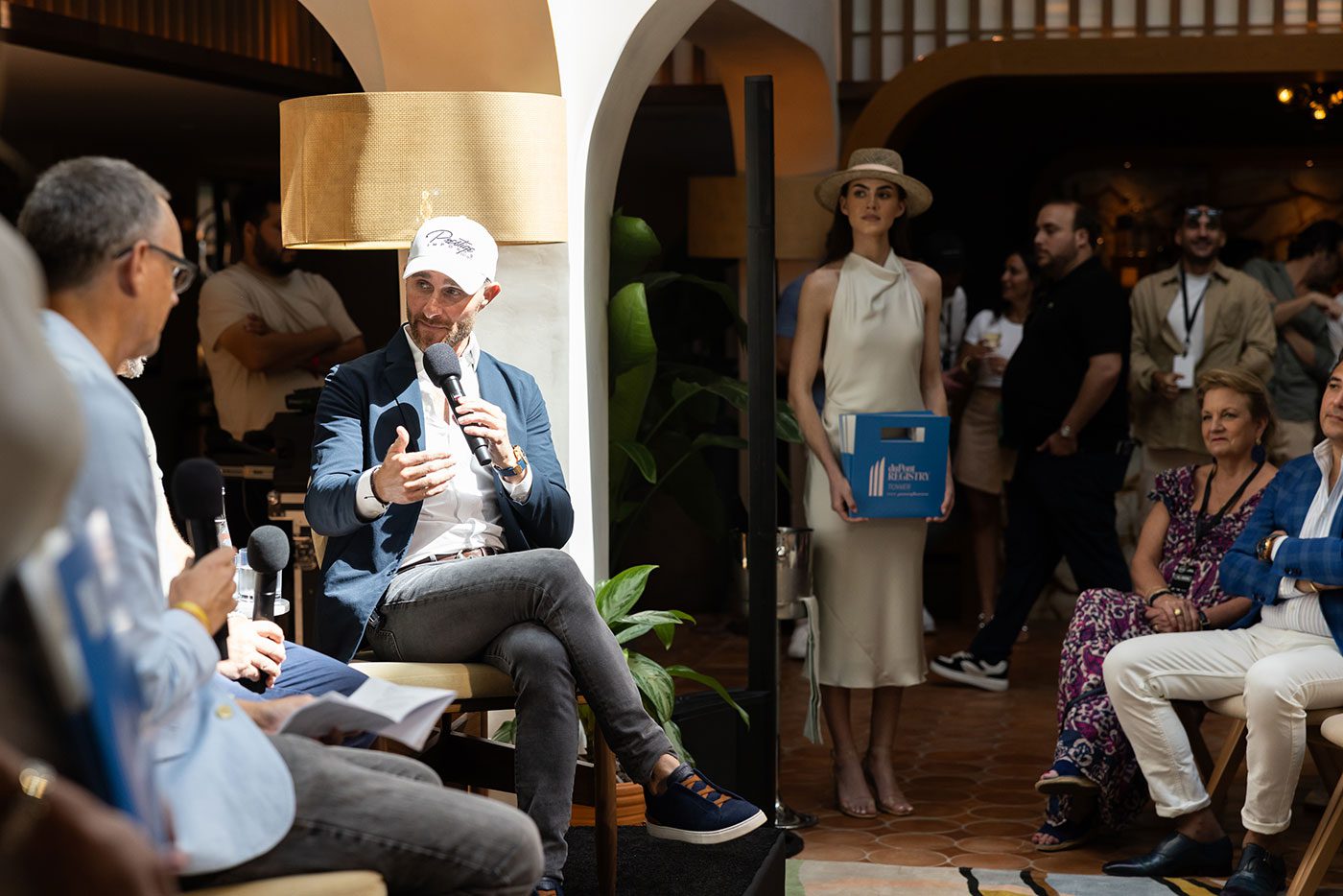 A man in a cap speaks into a microphone during an indoor event at the Miami Concours 2025, with an audience seated around him and a woman standing nearby holding a blue box, highlighting the essence of automotive luxury.