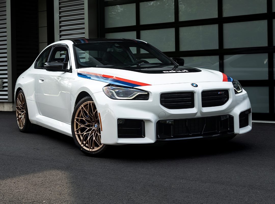 A white BMW 2002 Turbo with blue and red racing stripes and gold wheels is parked on black pavement in front of a modern building with glass doors, showcasing its classic Turbo Design Edition appeal.