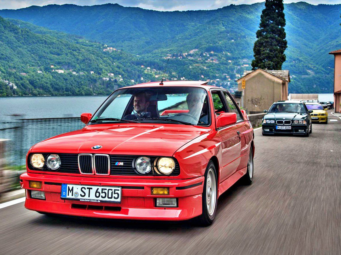 A red BMW E30 M3 drives along a lakeside road with mountains in the background, followed by another important car.