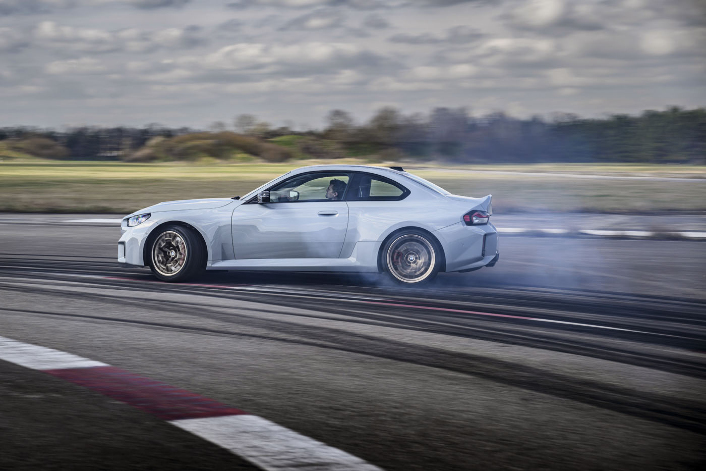 A white BMW 2026 M2 CS sports car drifting on a racetrack, with tire smoke and skid marks visible, under a cloudy sky—showcasing the power of its 523HP engine.
