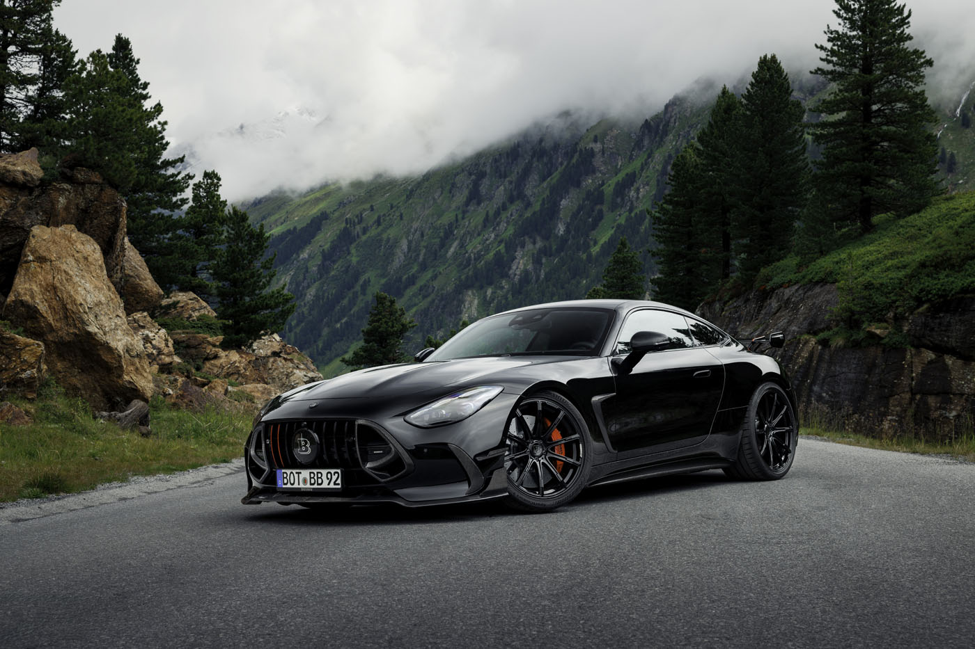 A BRABUS AMG GT hybrid supercar is parked on a mountain road, surrounded by trees and rocky cliffs with misty mountains in the background.