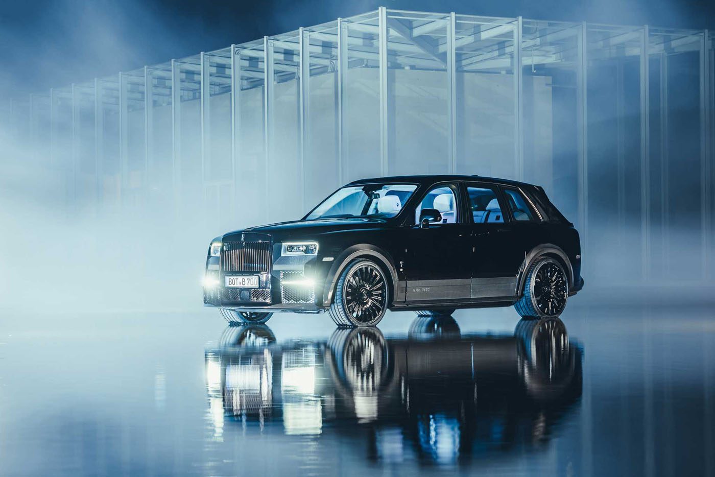 A black Rolls-Royce Cullinan is elegantly parked on a reflective surface in front of a modern architectural structure, enveloped by mist and dramatic lighting.