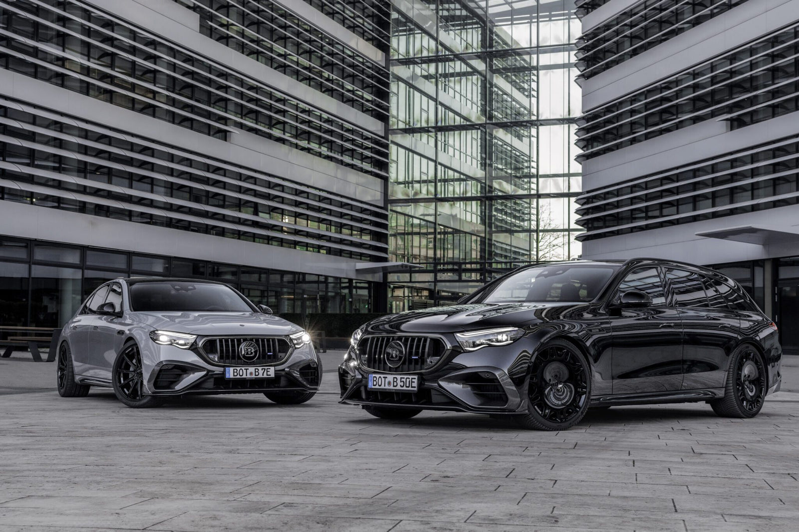 Two Mercedes-AMG E53 cars, a sedan and a wagon, are parked side by side in a modern urban courtyard with glass and steel buildings in the background, highlighting the possibilities of the AMG personalization program.