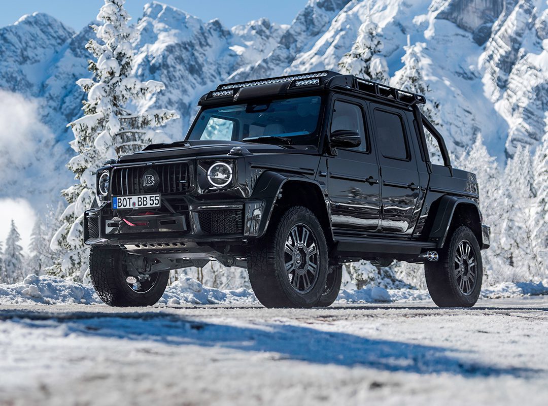 A black BRABUS XLP 800 ADVENTURE pickup truck, based on the Mercedes-AMG G63, is parked on a snowy mountain road with snow-covered trees and mountains in the background.