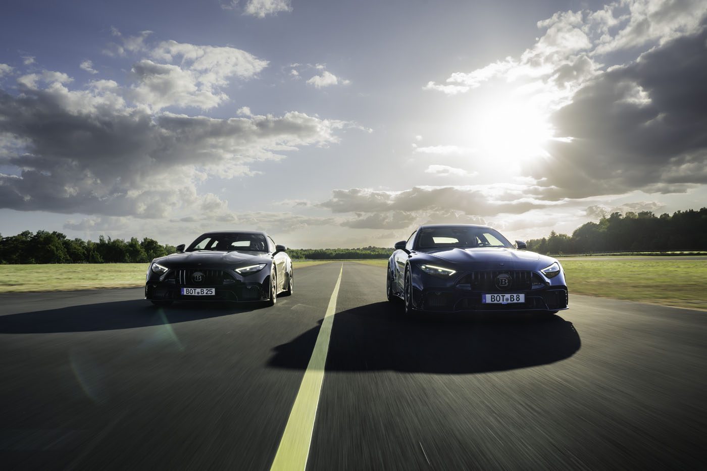 Two black sports cars drive side by side on an empty road under a partly cloudy sky with the sun shining in the background.