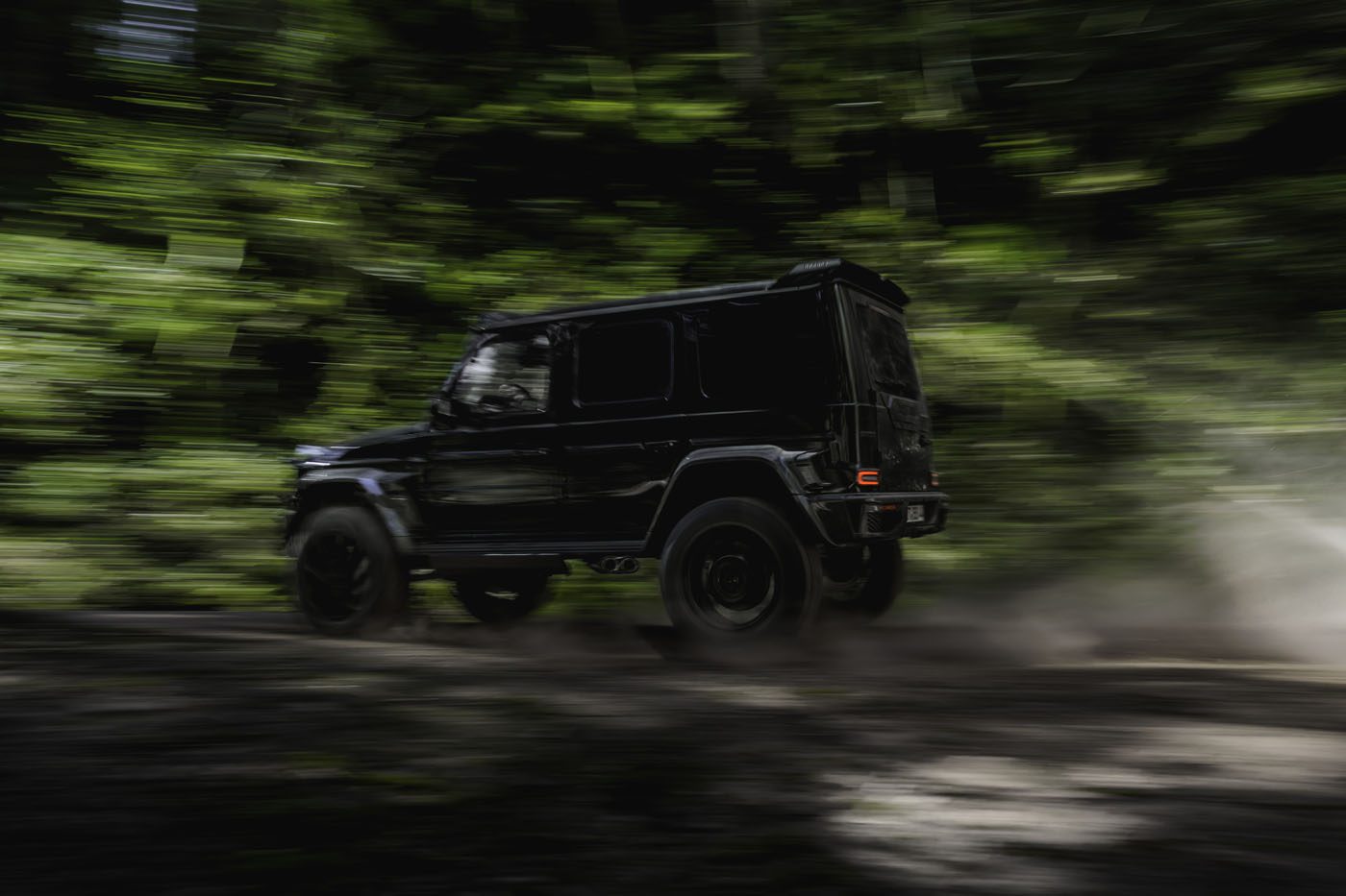A BRABUS G 63 XL 800 Off-Roader speeds along a dirt road through a lush, green forest, with motion blur emphasizing the SUV’s power and agility.