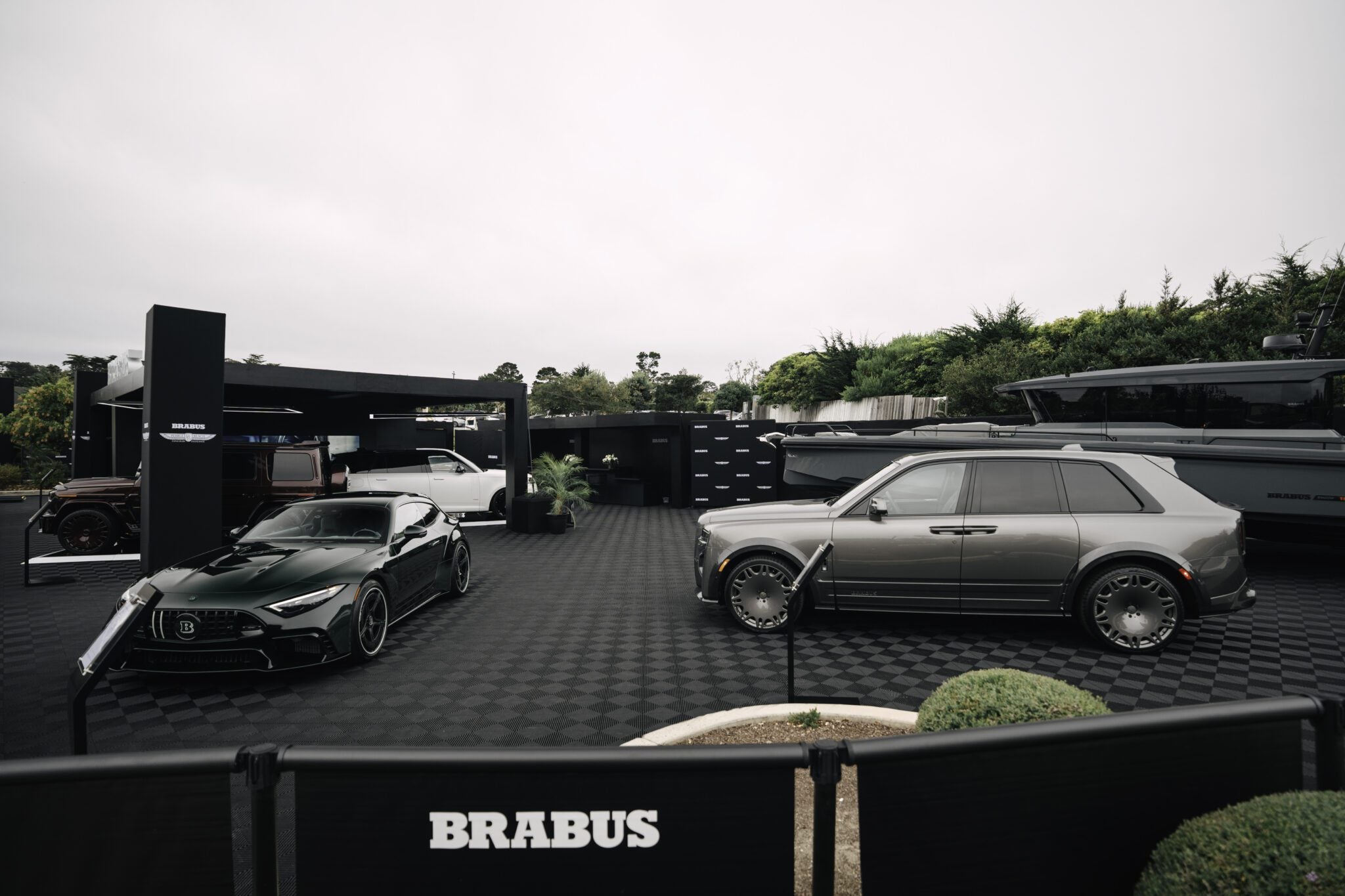 Two luxury cars—a dark green sports car and a silver SUV—are showcased in the outdoor BRABUS exhibition area at Pebble Beach, with a yacht and lush greenery completing the stylish backdrop.